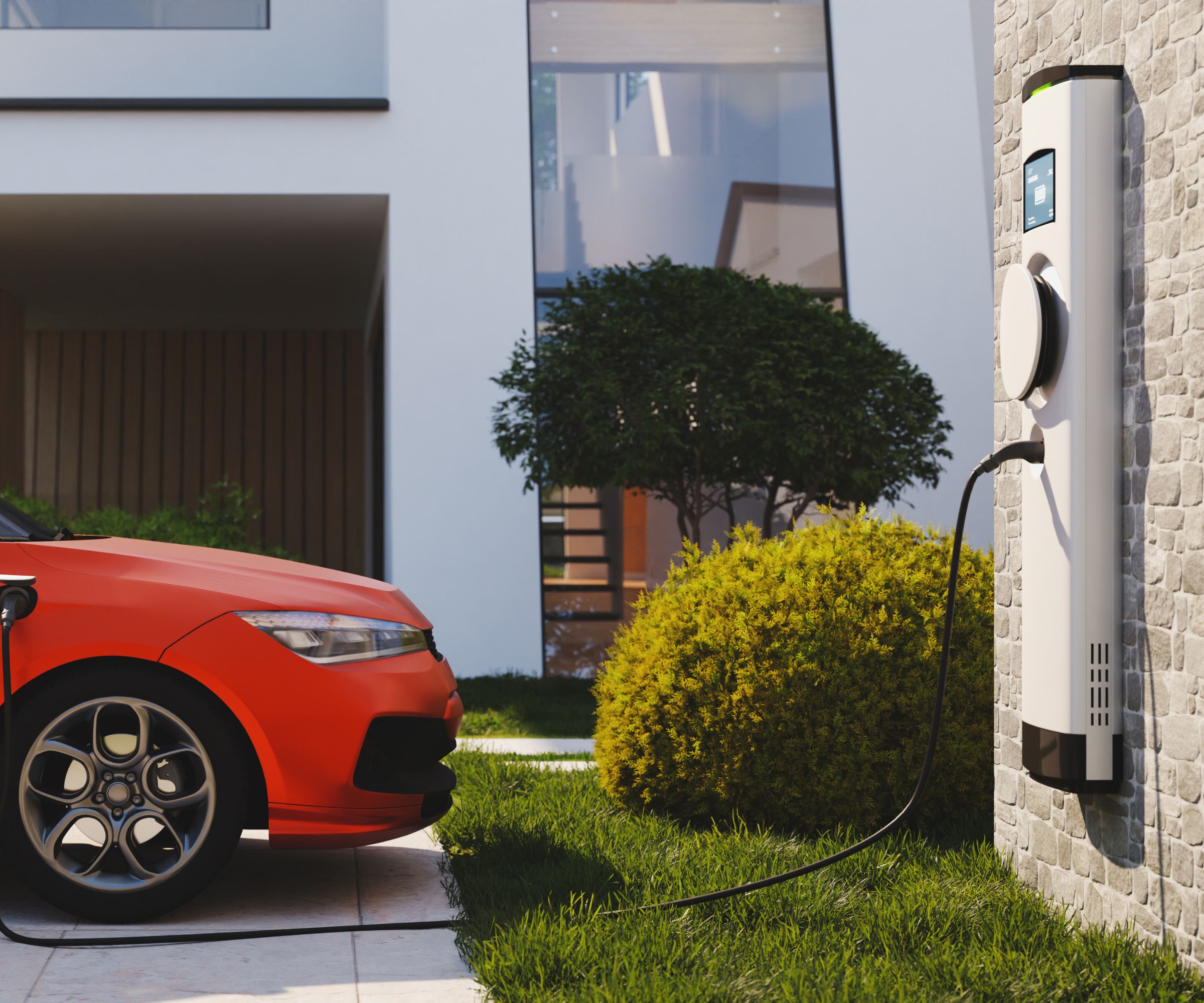 Une borne de recharge pour voiture électrique fixée au mur d'une maison rechargeant une voiture rouge dans une allée. Une maison moderne en arrière-plan